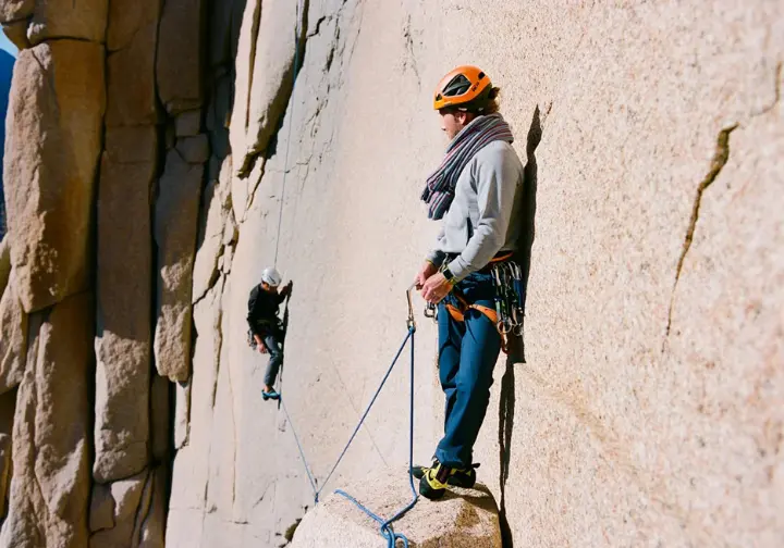 A climber belays their partner from above at an intermediate station on a sunny cliff, clearly illustrating the definition of multi-pitch climbing.