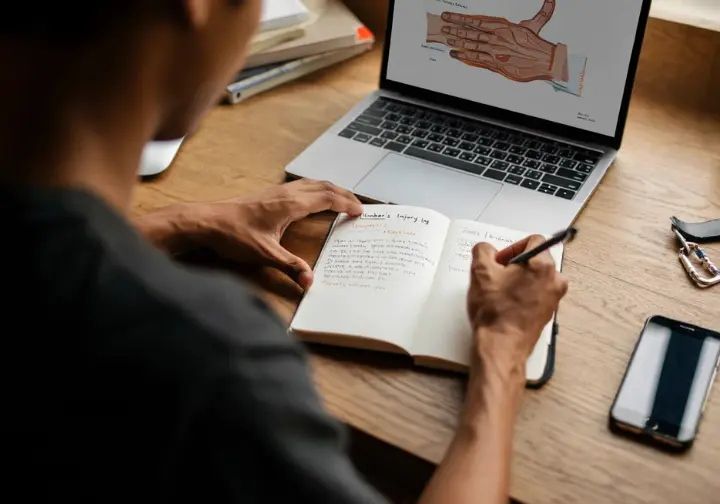 A climber preparing for a doctor's appointment by writing in an injury log at their desk, with a laptop showing hand anatomy nearby.