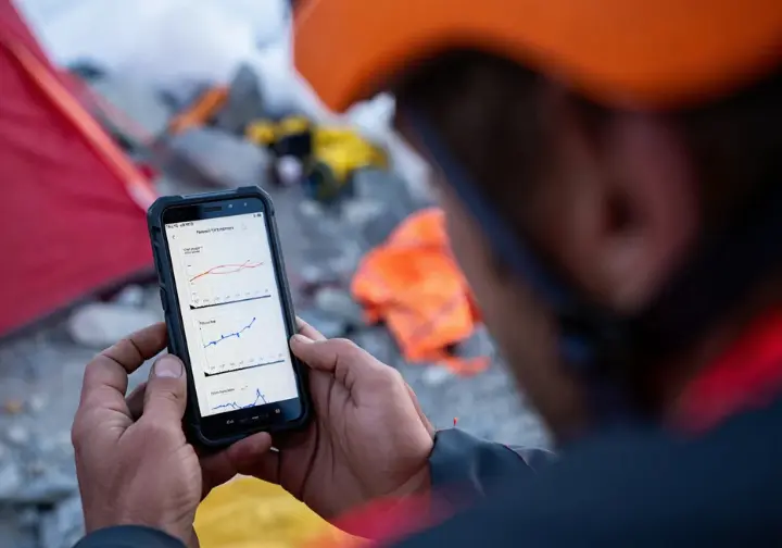 A close-up of a climber's hands holding a smartphone, which displays a detailed hourly weather graph for a mountain forecast.