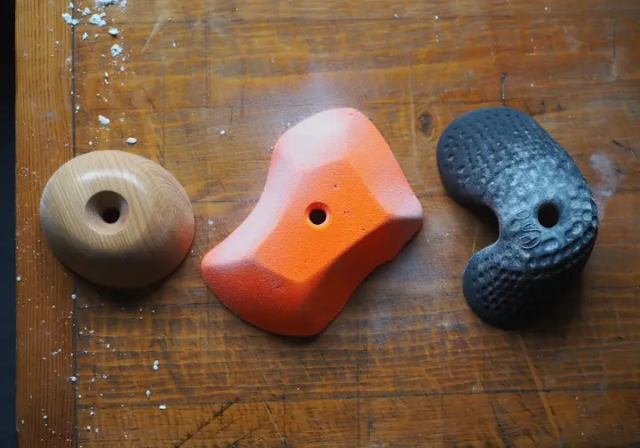 A detailed comparison of three climbing holds on a workbench: a wooden Tension hold, a plastic Kilter hold, and a plastic MoonBoard hold.