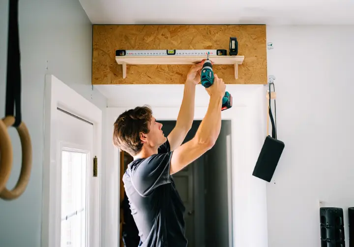 A person installing a wooden hangboard with a power drill, building their own rock climbing training setup in their home.