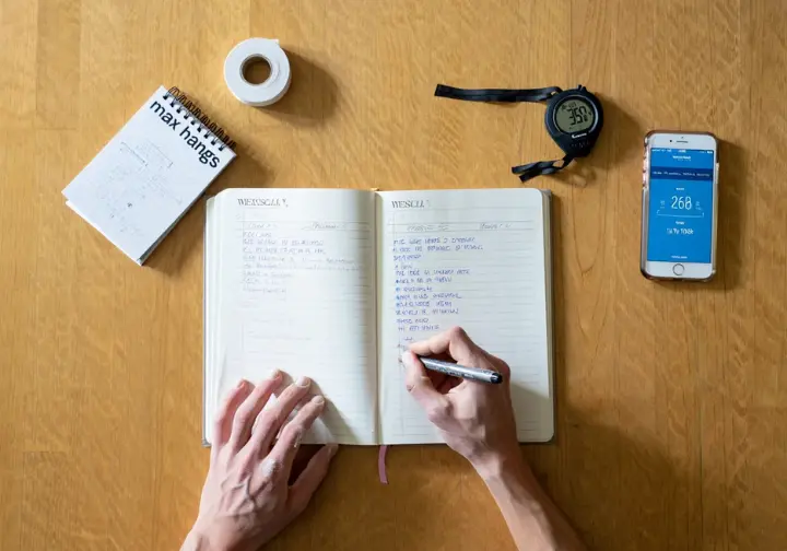A top-down view of a climber's hands writing in a training journal, surrounded by tools like a stopwatch and finger tape, showing the step-by-step process of building a plan.