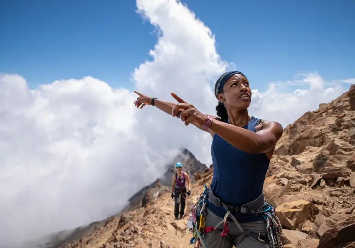 A climber on a mountain trail points up to the sky, showing her partner how to interpret the vertical growth of cumulus clouds as a sign of changing weather.