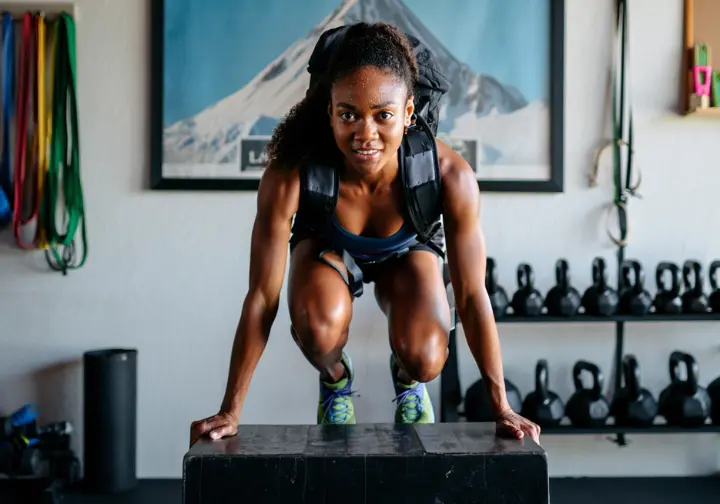 A female climber in a home gym performs weighted step-ups with a backpack on, diligently following a preparation protocol for mountaineering.