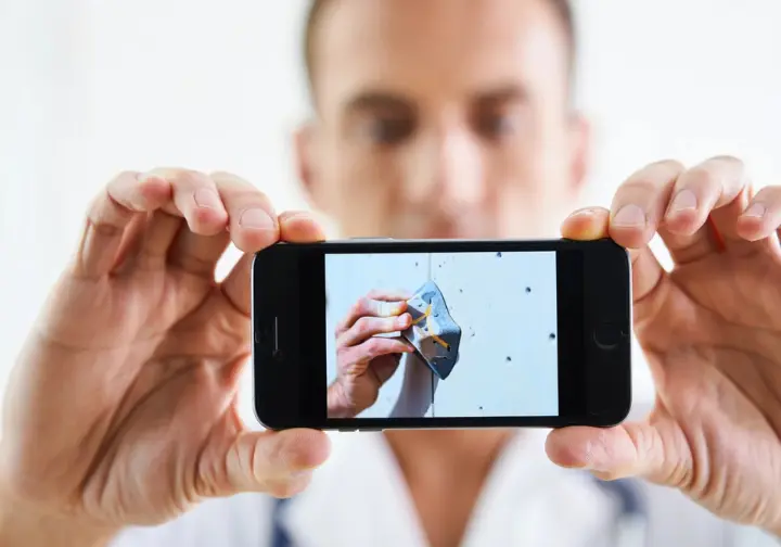 A close-up of a climber's hands showing their doctor a photo of a specific climbing hold on a smartphone to explain their injury mechanism.