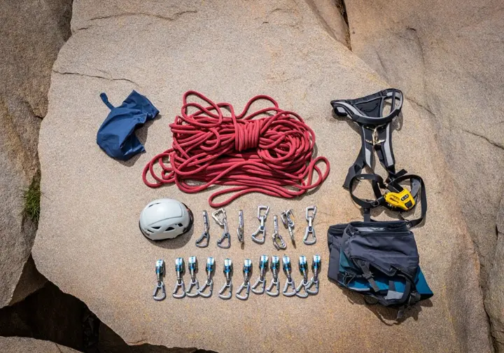 A complete set of essential multi-pitch climbing gear, including a rope, trad rack, harness, and helmet, neatly organized on a flat rock at a crag.