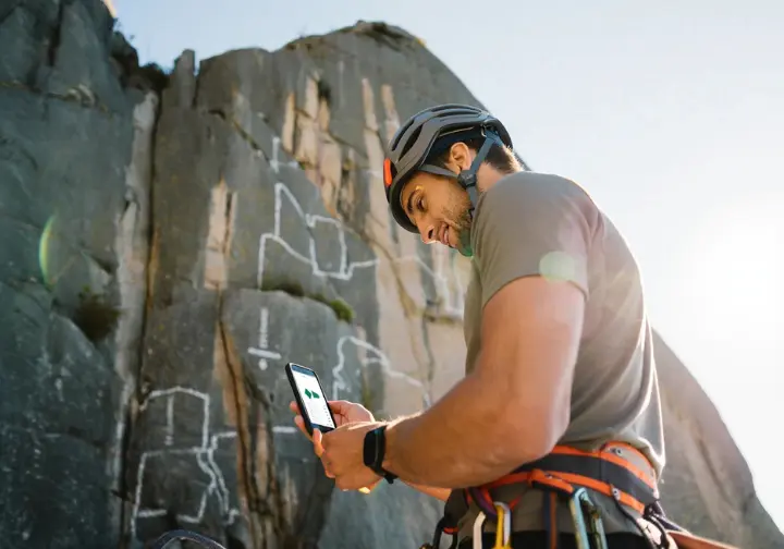 A geared-up climber at the base of a cliff reviews his daily training plan on his smartphone before starting his climb, putting his plan into practice.