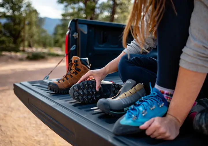 A climber performs a comparative analysis, examining a hiking boot, approach shoe, and climbing shoe laid out on a truck's tailgate.