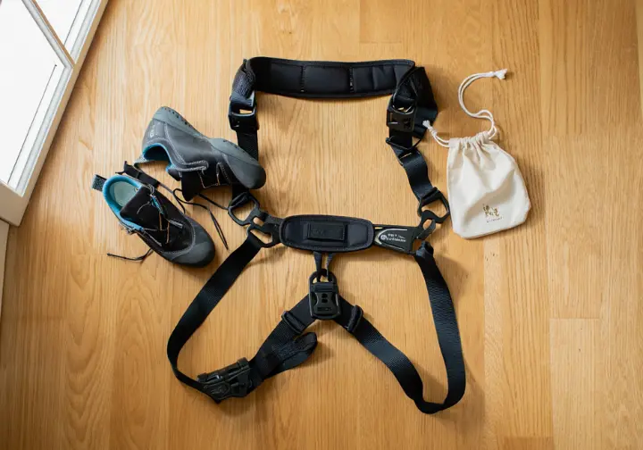 A flat lay of essential pregnancy climbing gear, featuring a Mad Rock Mountain Mama full-body harness, climbing shoes, and a chalk bag.
