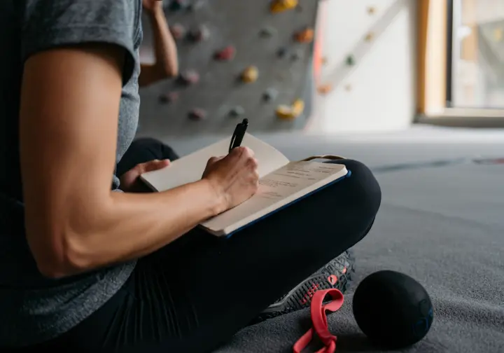 A climber sits in a gym writing in a training journal, planning their personalized mobility program with tools nearby.