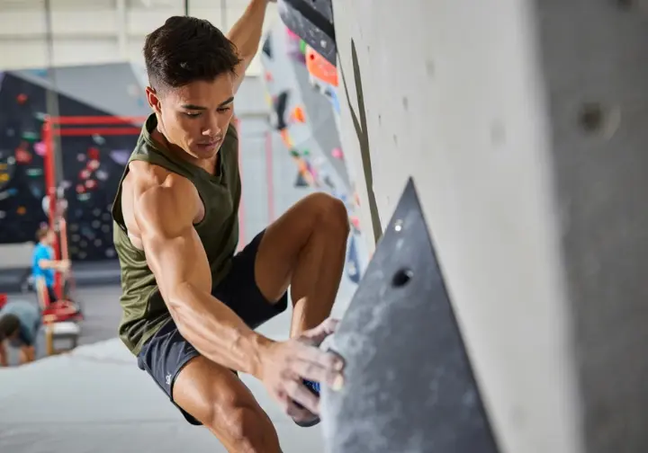 A climber in a gym demonstrates a progressive training framework by engaging in a quiet feet drill, focusing intently on precise foot placement.