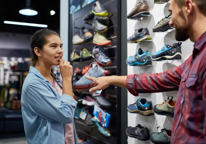 A climber gets expert advice from a store employee while looking at a wall of hiking and climbing shoes in a buying guide scenario.