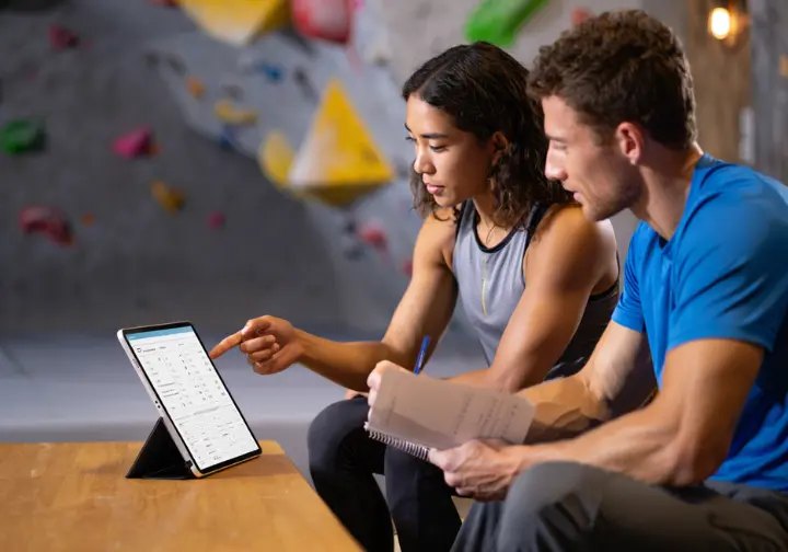 Two climbers work together to create a personalized rock climbing training plan using a tablet and a journal in a gym.