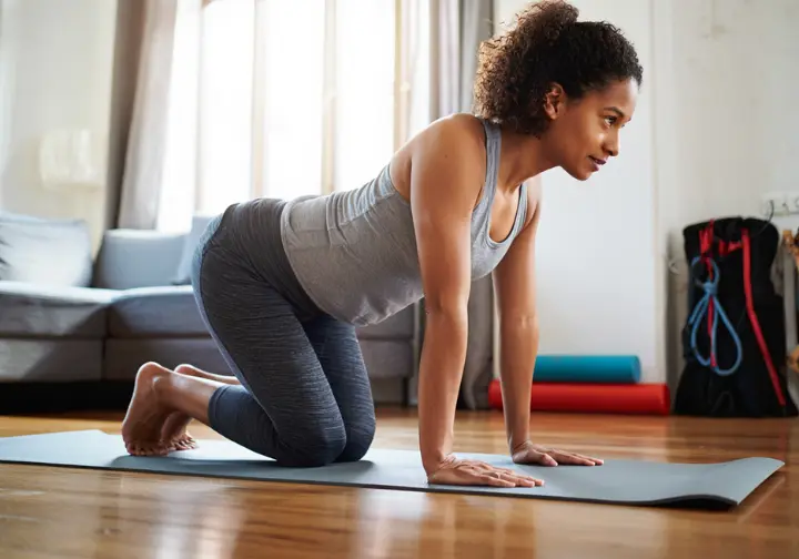 A new mother performs gentle core exercises on a yoga mat at home, with her climbing gear in the background, showing her postpartum return to climbing.