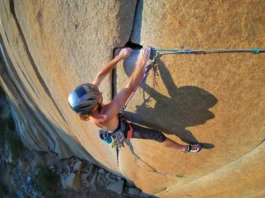 A climber in full aid gear, including a helmet and harness, stands in aiders high on a sheer granite cliff, reaching up to place protection.