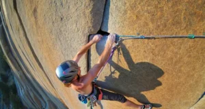 Aid Climbing Guide: A0-A5 Grades & Gear Explained A climber in full aid gear, including a helmet and harness, stands in aiders high on a sheer granite cliff, reaching up to place protection.