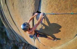 Aid Climbing Guide: A0-A5 Grades & Gear Explained A climber in full aid gear, including a helmet and harness, stands in aiders high on a sheer granite cliff, reaching up to place protection.