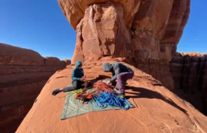 Arches National Park Climbing Rules 2025: Live Closure Atlas Two climbers in full outdoor gear organize their ropes and equipment at the base of a towering red sandstone formation in Arches National Park.