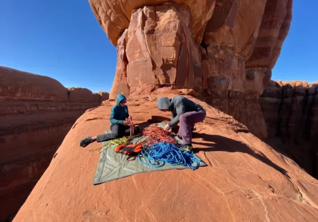 Two climbers in full outdoor gear organize their ropes and equipment at the base of a towering red sandstone formation in Arches National Park.