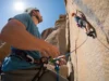 Belay Devices Decoded: ATC, GriGri & Passive Explained A belayer in a full-body shot stands on the ground, carefully managing the rope for a climber on a sunny rock face above.