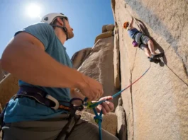 A belayer in a full-body shot stands on the ground, carefully managing the rope for a climber on a sunny rock face above.