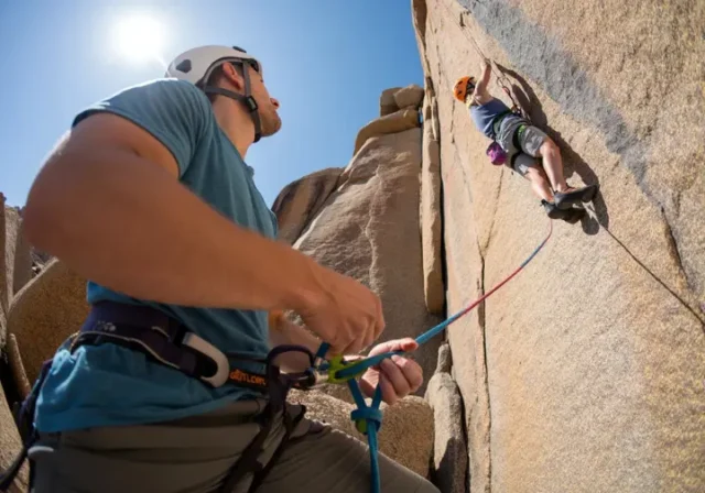 A belayer in a full-body shot stands on the ground, carefully managing the rope for a climber on a sunny rock face above.