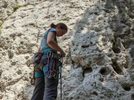 A full-body shot of a female climber in pants and t-shirt standing at the bottom of a rock cliff, looking down at the quickdraws on her harness.