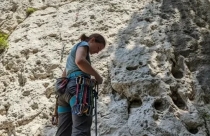 How to Choose Your First Quickdraws: 2025 Expert Guide A full-body shot of a female climber in pants and t-shirt standing at the bottom of a rock cliff, looking down at the quickdraws on her harness.