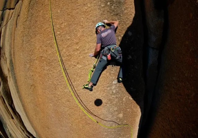 A full-body shot of a climber in a helmet leading a sport climb on a sunny day, with the climbing rope visible below.