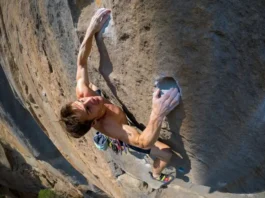 A full-body shot of a rock climber in the middle of a difficult move on a sunny, overhanging cliff, showcasing the concept of power-to-weight ratio.
