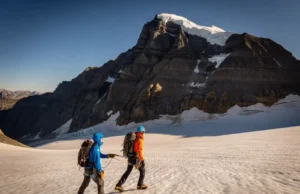 Mount Robson Emperor Face Guide: M7 Rack & Speed Plan Full-body shot of two alpinists in full climbing gear walking across a glacier, with the enormous Emperor Face of Mount Robson looming behind them.