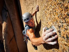 Full-body shot of a trad climber in a helmet and harness placing a cam in a sunlit rock crack, as seen from a low angle.