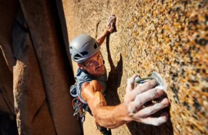 Trad Protection 102: The Ultimate Cam Guide for All Climbers Full-body shot of a trad climber in a helmet and harness placing a cam in a sunlit rock crack, as seen from a low angle.