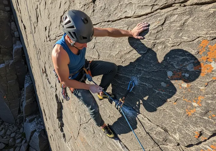 A full-body shot of a climber on a rock face, looking down at a nut they have just placed for protection.