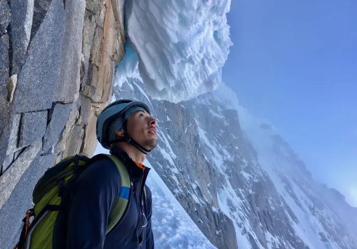 A lone alpinist in a full-body shot stands dwarfed at the bottom of the Emperor Face, looking up at the immense and hazardous ice-covered cliff.
