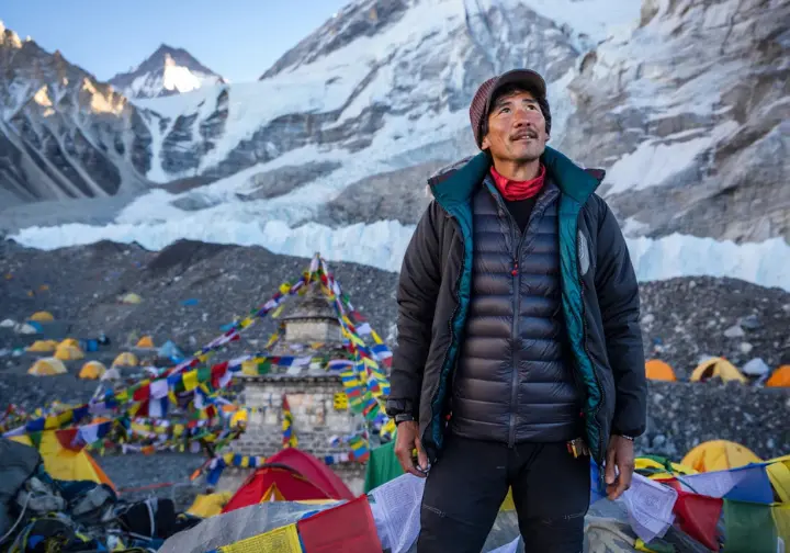 A climber in a full-body shot stands at Everest Base Camp, looking up at the Khumbu Icefall with expedition tents in the background.