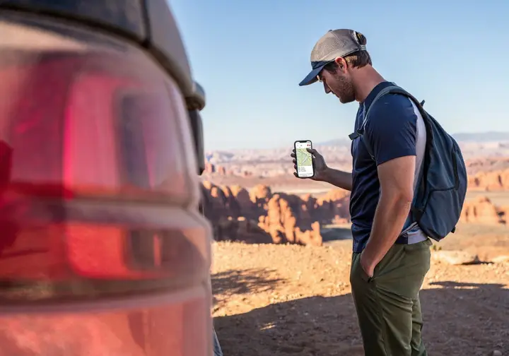 A climber in hiking clothes stands by their car, looking at a map on their smartphone to plan a route in Arches National Park.