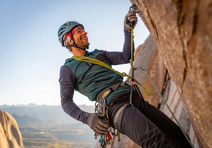 A climber in a helmet and full gear stands at a belay anchor high on a cliff, using an advanced belay technique to protect their partner.