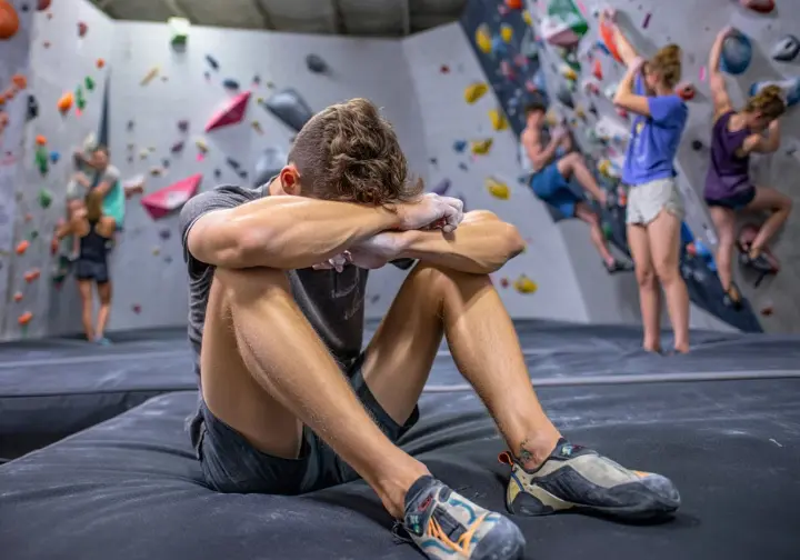 A full-body shot of an exhausted climber slumping on a crash pad in a climbing gym, representing the metabolic cost and energy depletion from a zero-carb diet.