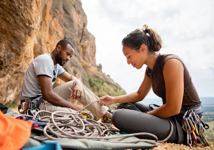 Two climbers in a full-body shot sitting at the base of a cliff, organizing a new set of climbing nuts on their gear.