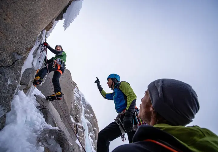 Two alpinists in full gear stand at a belay ledge on an ice wall, with one pointing up to discuss the path ahead on the Emperor Face.