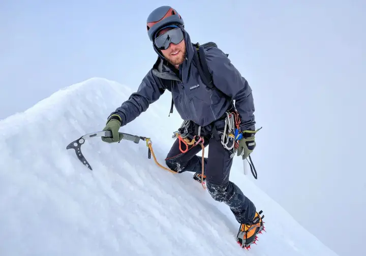 A full-body shot of a mountaineer using an ice axe in the dagger position to climb a steep snow couloir.