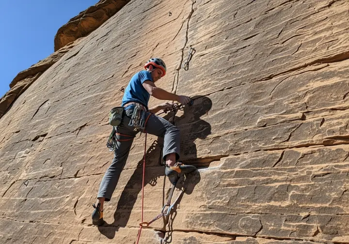 View from below of a climber in a full-body shot on a steep rock face, assessing a crack to make a cam placement.