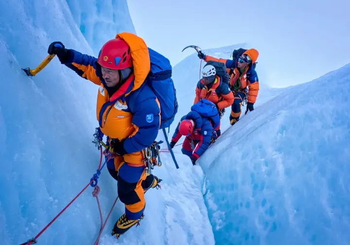 Full-body shot of three climbers in down suits ascending the steep, icy Lhotse Face on Mount Everest using fixed ropes.