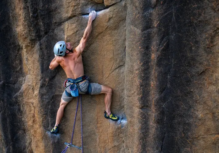 A climber in a full-body shot demonstrates the workflow of aid climbing, standing high in an aider to place the next piece of gear on a cliff.