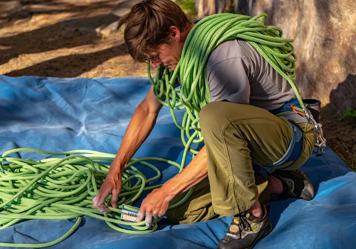 A full-body shot of a climber wearing climbing pants and a t-shirt, carefully flaking a new rope onto a tarp at the base of a cliff.