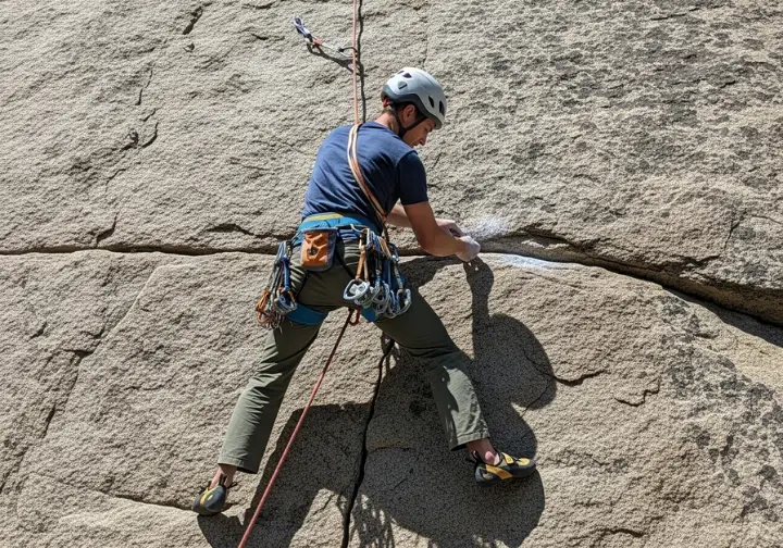 A climber in a full-body shot is practicing placing trad gear on a rock face while safely secured by a toprope.