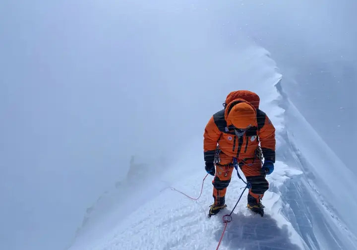 A lone climber in a full-body shot battles high winds and blowing snow on an exposed ridge high on Mount Everest.
