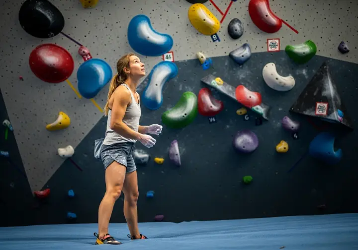 A full-body shot of a confident female climber looking up at a bouldering wall, representing the progression after a training plan.