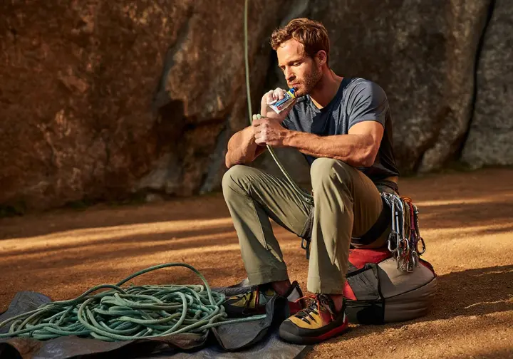 A climber in full gear sits at the base of a crag, about to consume an energy gel before their climb, illustrating a smart, targeted carbohydrate strategy.